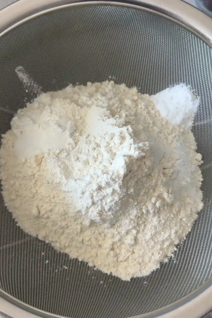 A fine mesh sieve filled with flour and dry ingredients like baking soda and salt, sitting over a bowl as the ingredients are being sifted together.