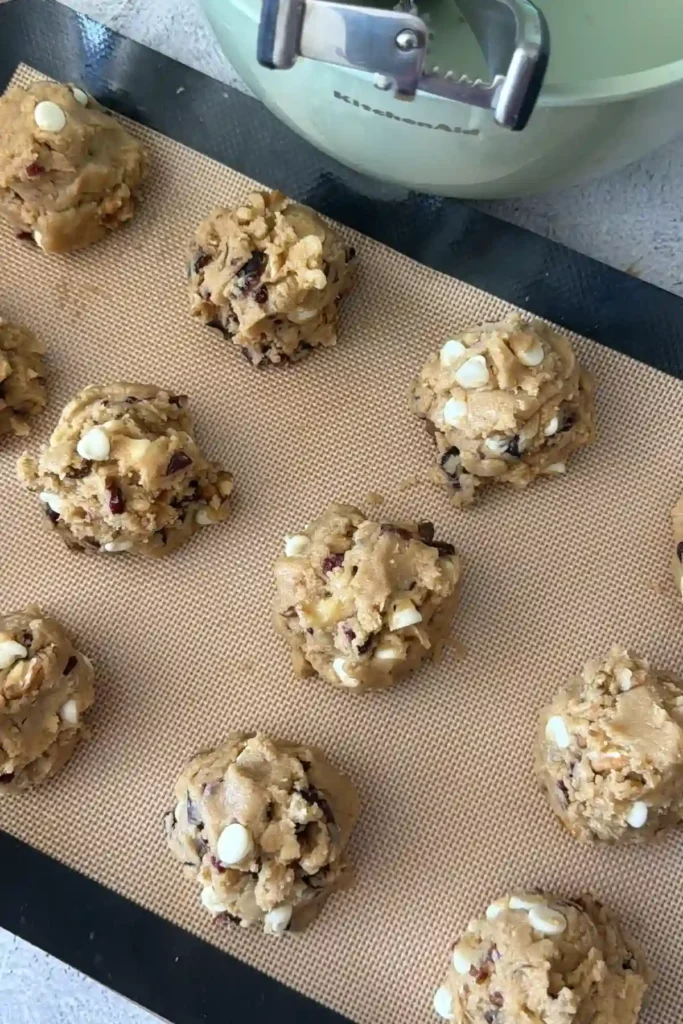 Scoops of cookie dough placed on a silicone-lined baking sheet, spaced apart and ready for baking, with a cookie scoop visible in the background.