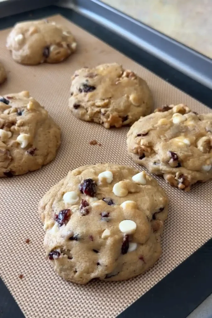 Freshly baked cookies on a silicone mat, slightly spread with golden edges and visible chunks of white chocolate and cranberries.