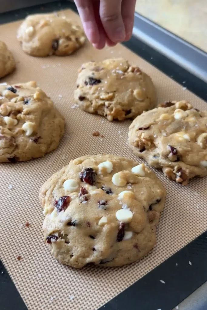 A close-up of baked cookies on a tray as flaky sea salt is being sprinkled on top, highlighting texture and finishing touch.