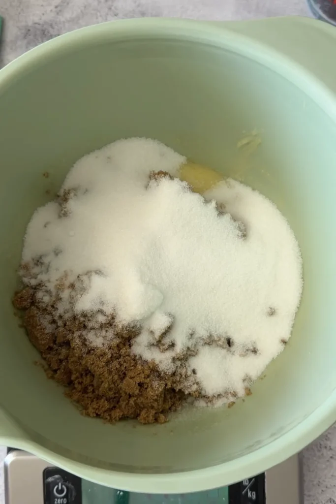 A mixing bowl on a kitchen scale with brown sugar and white sugar piled on top of softened butter, ready to be creamed together.