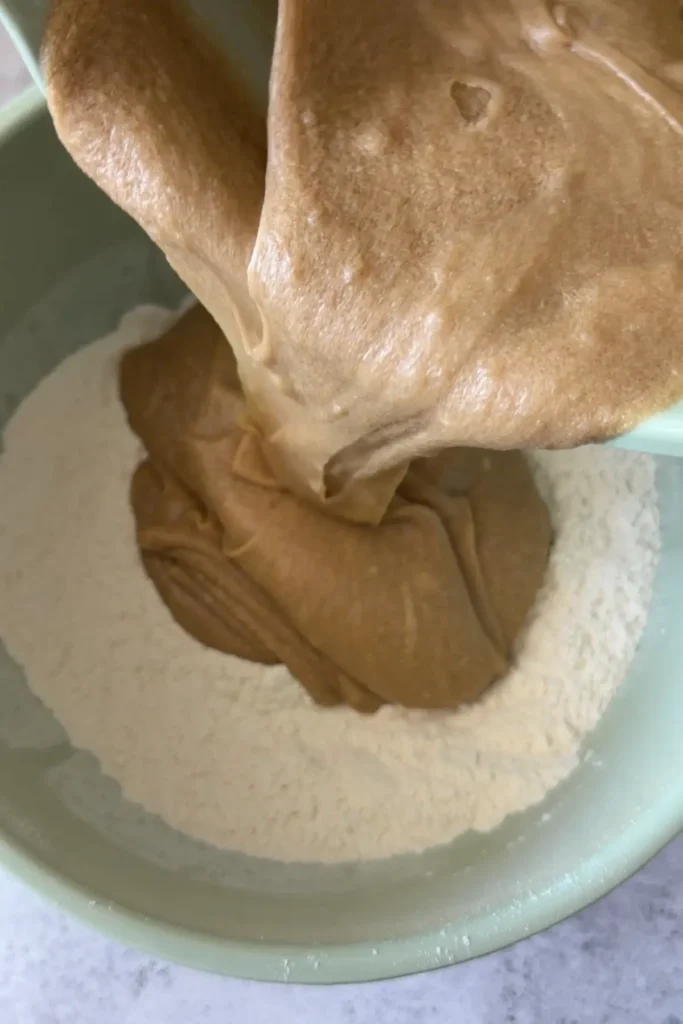The wet mixture being poured into the bowl of dry ingredients, creating a contrast between the light flour and darker batter.