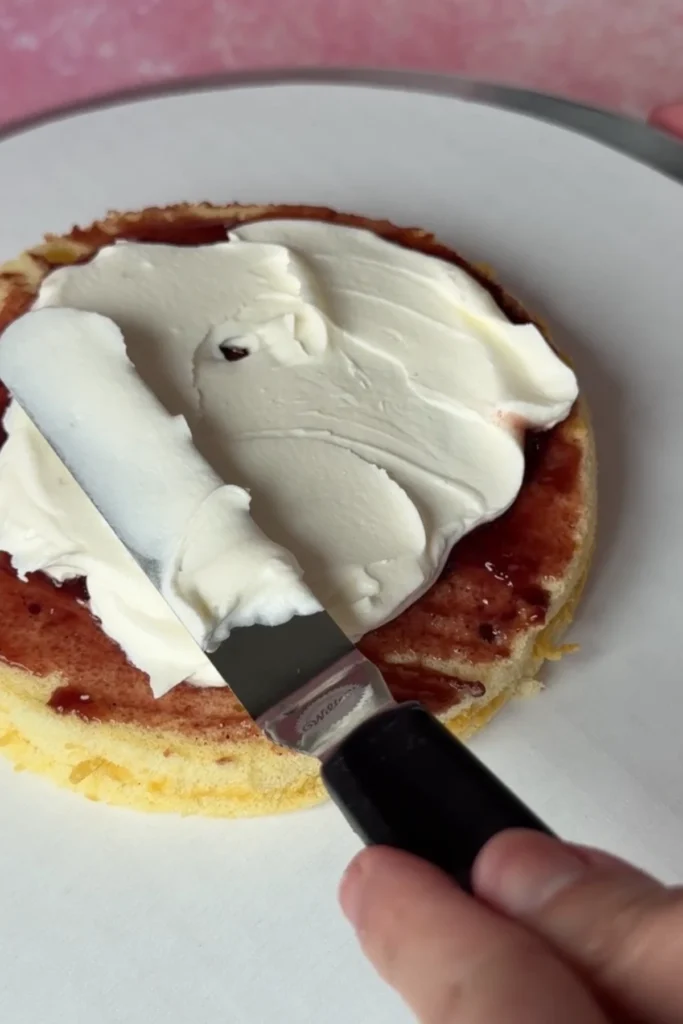 Offset spatula spreading mascarpone chantilly cream over berry jam on a chiffon cake layer during assembly.