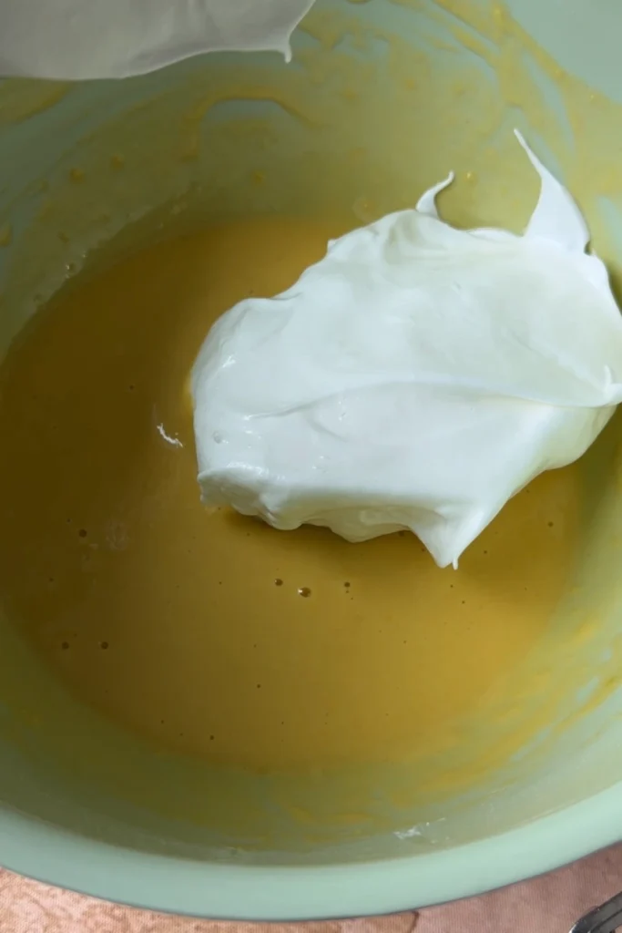 Whipped egg whites being added into a pale yellow egg yolk batter during the sponge cake mixing process.