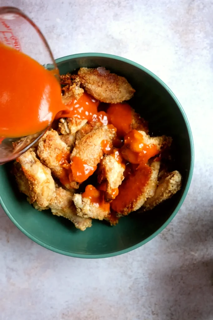 Homemade buffalo sauce being poured over crispy air fryer chicken wings in a green mixing bowl.