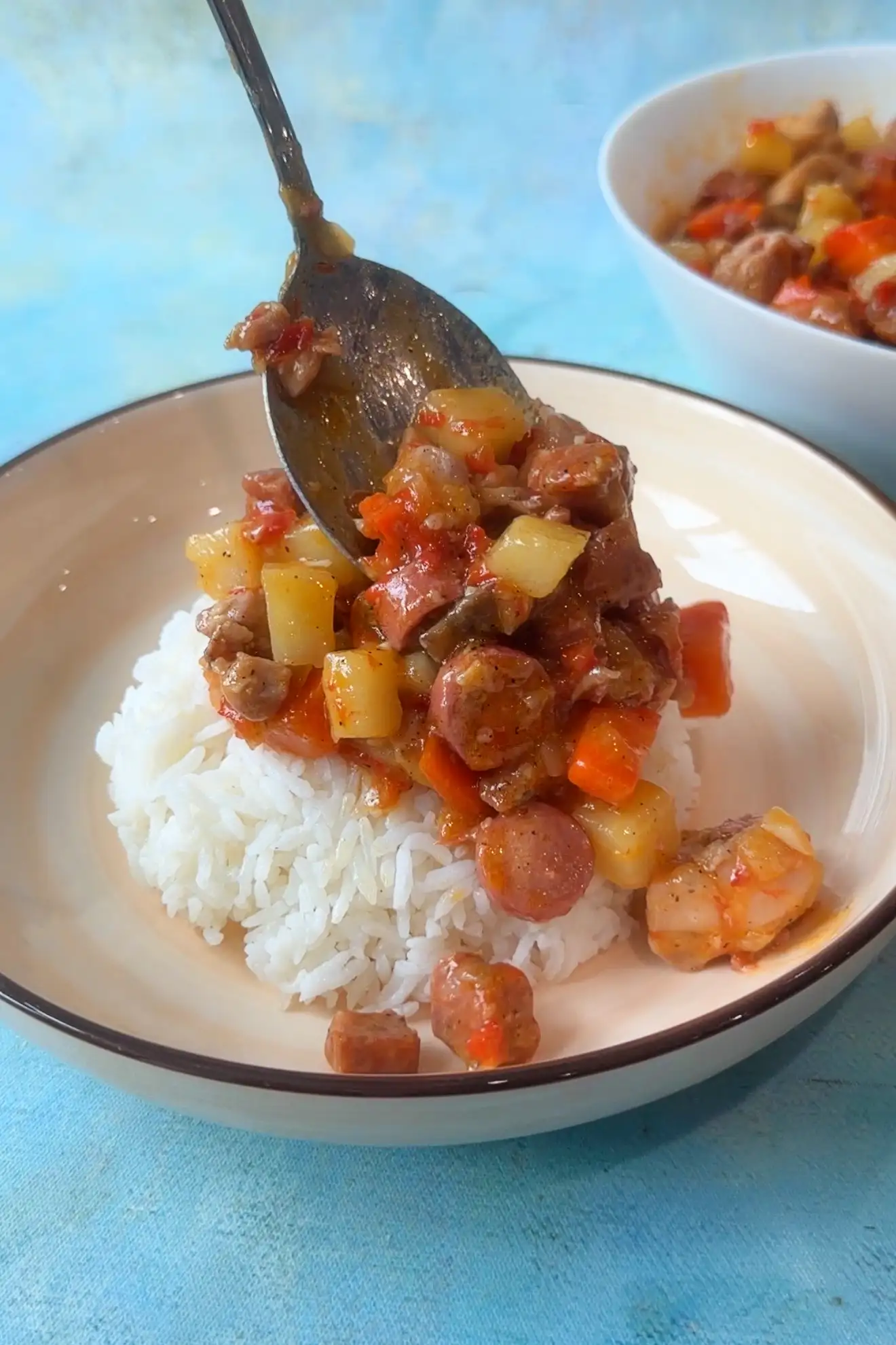 Spoon serving chicken pastel over steamed rice, featuring chicken, potatoes, carrots, mushrooms, and sausage.