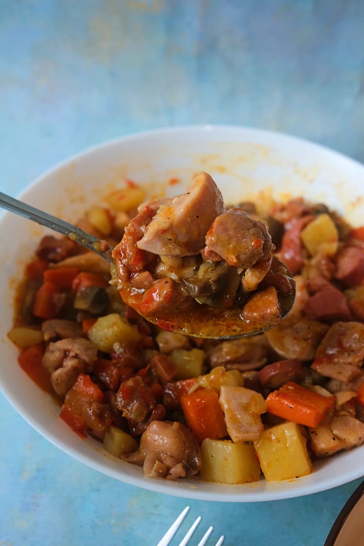 Spoonful of chicken pastel lifted from a bowl, showing tender chicken, mushrooms, potatoes, carrots, and sausage.