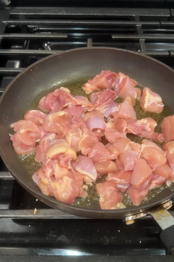 Raw chicken thigh pieces cooking in oil in a skillet on the stovetop at the start of making Filipino chicken pastel.