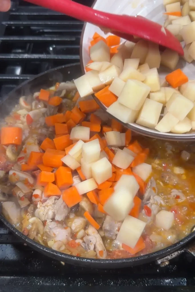 Diced potatoes and carrots being added to simmering chicken pastel in a skillet.