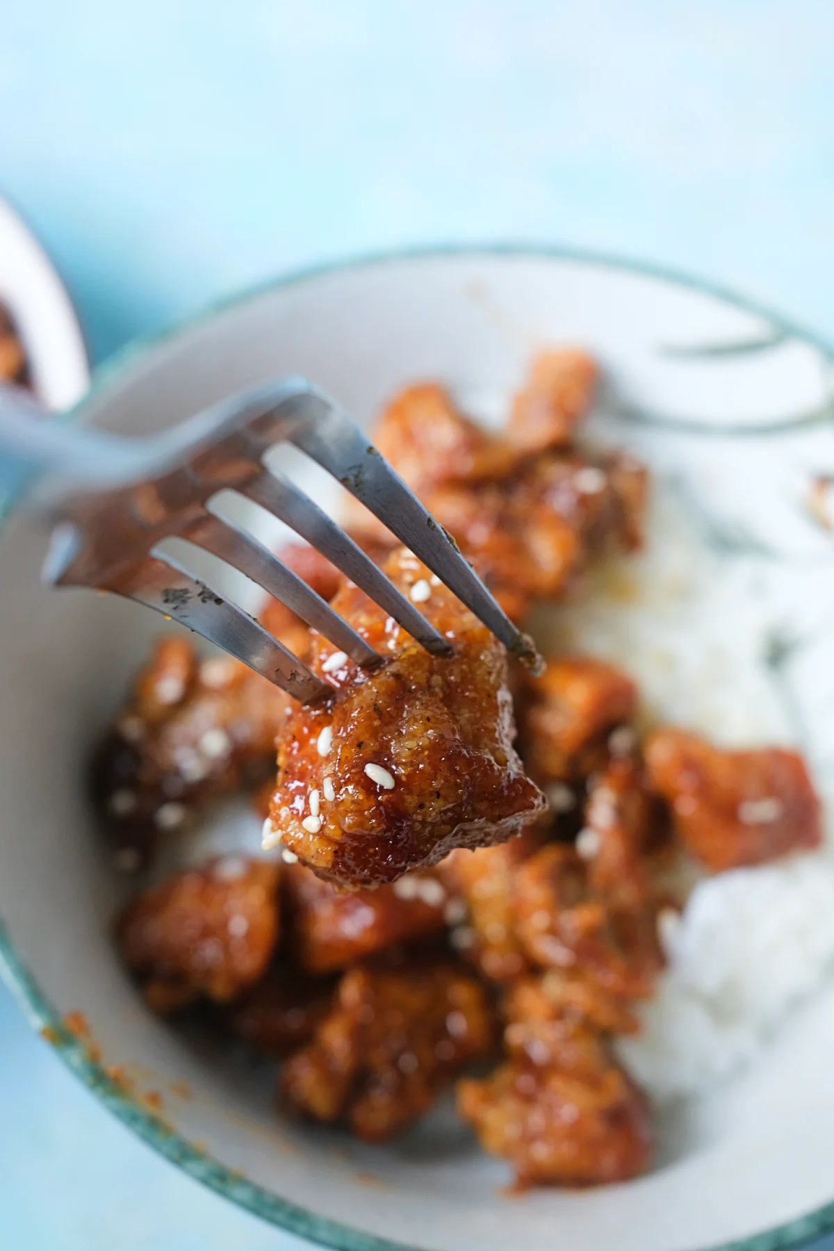 Fork holding a crispy piece of honey garlic chicken over a bowl of rice and chicken.