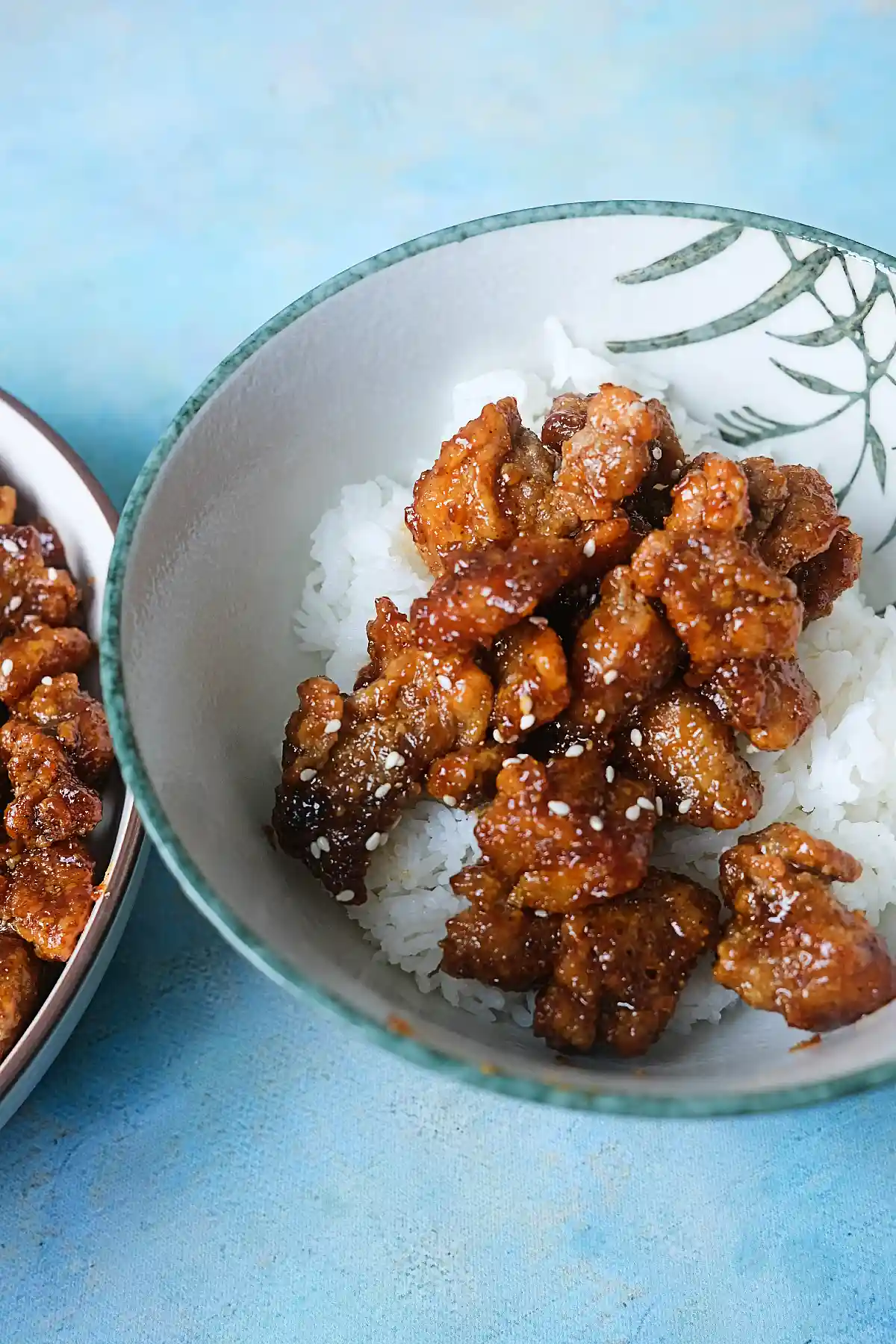 Crispy honey garlic chicken served over rice with sesame seeds on a light blue background.