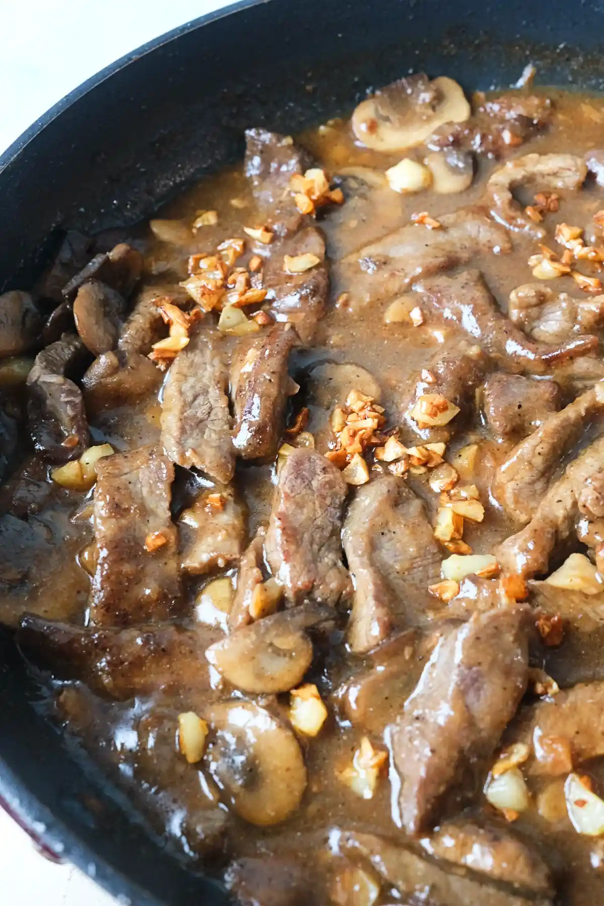 Garlic pepper beef with mushrooms cooking in a skillet with thick savory sauce.