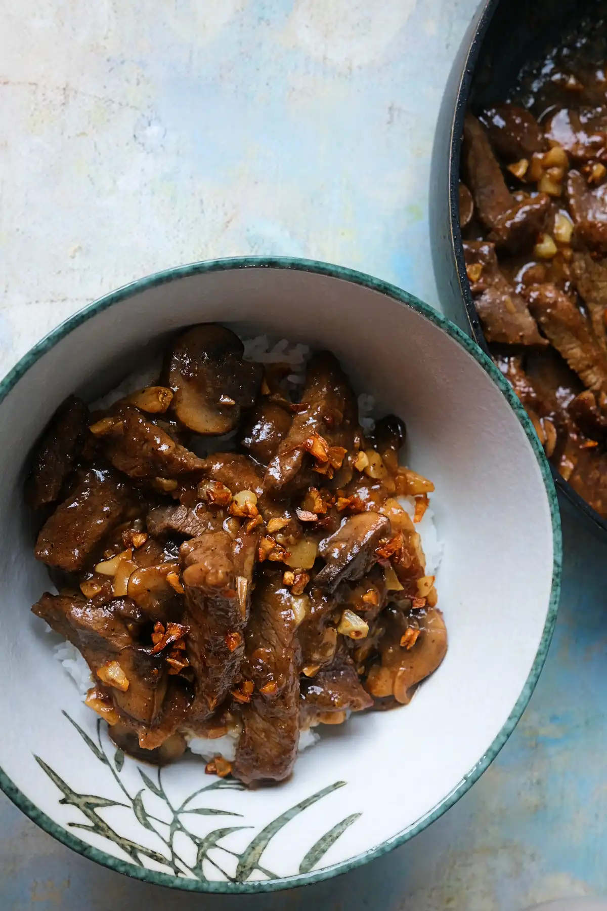 Overhead shot of mushroom pepper steak served over rice beside a skillet of garlic pepper beef.