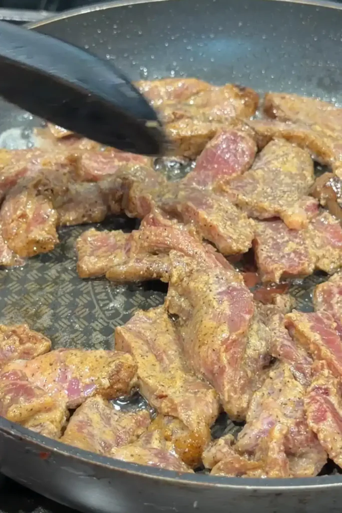 Marinated beef strips cooking in a hot skillet before browning.