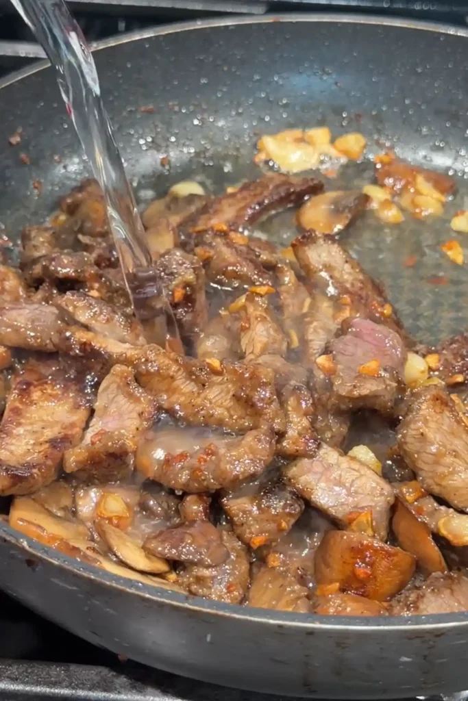 Water being poured into browned garlic pepper beef and mushrooms to create a savory pan sauce.
