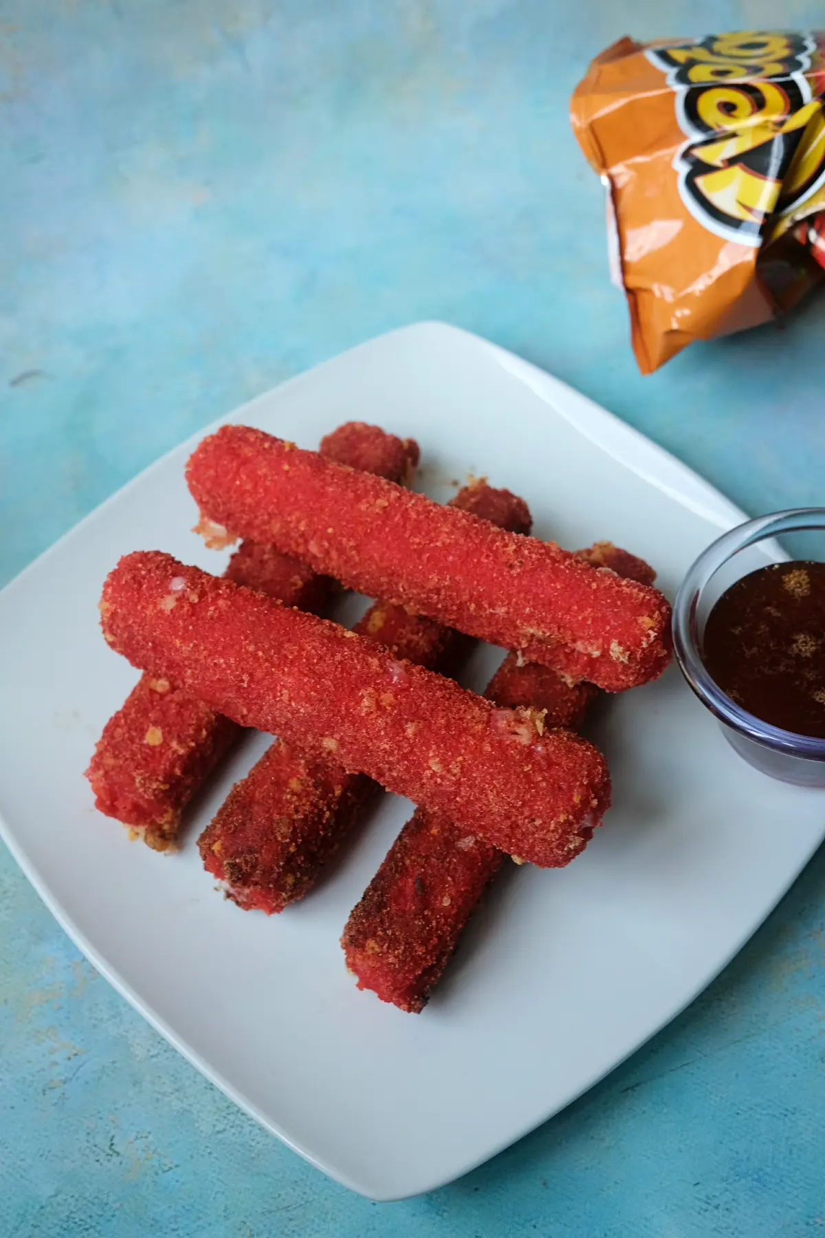 Homemade Hot Cheetos mozzarella sticks with hot honey dip, honey bottle, and Flamin’ Hot Cheetos bag in the background.