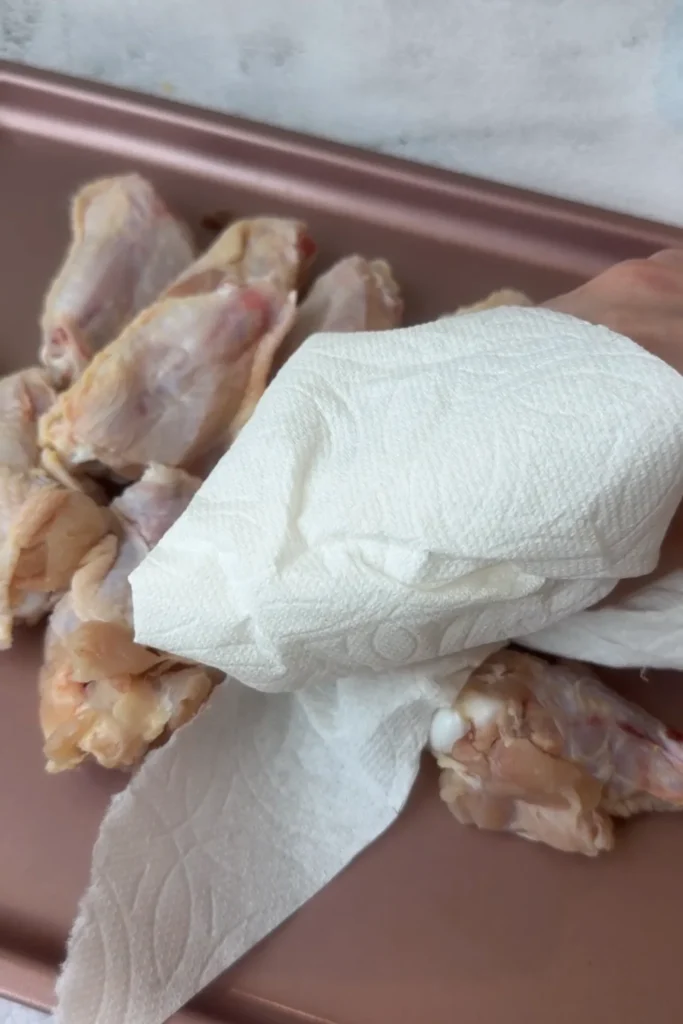 Patting raw chicken wings dry with paper towels on a baking tray before seasoning for crispy air fryer wings.