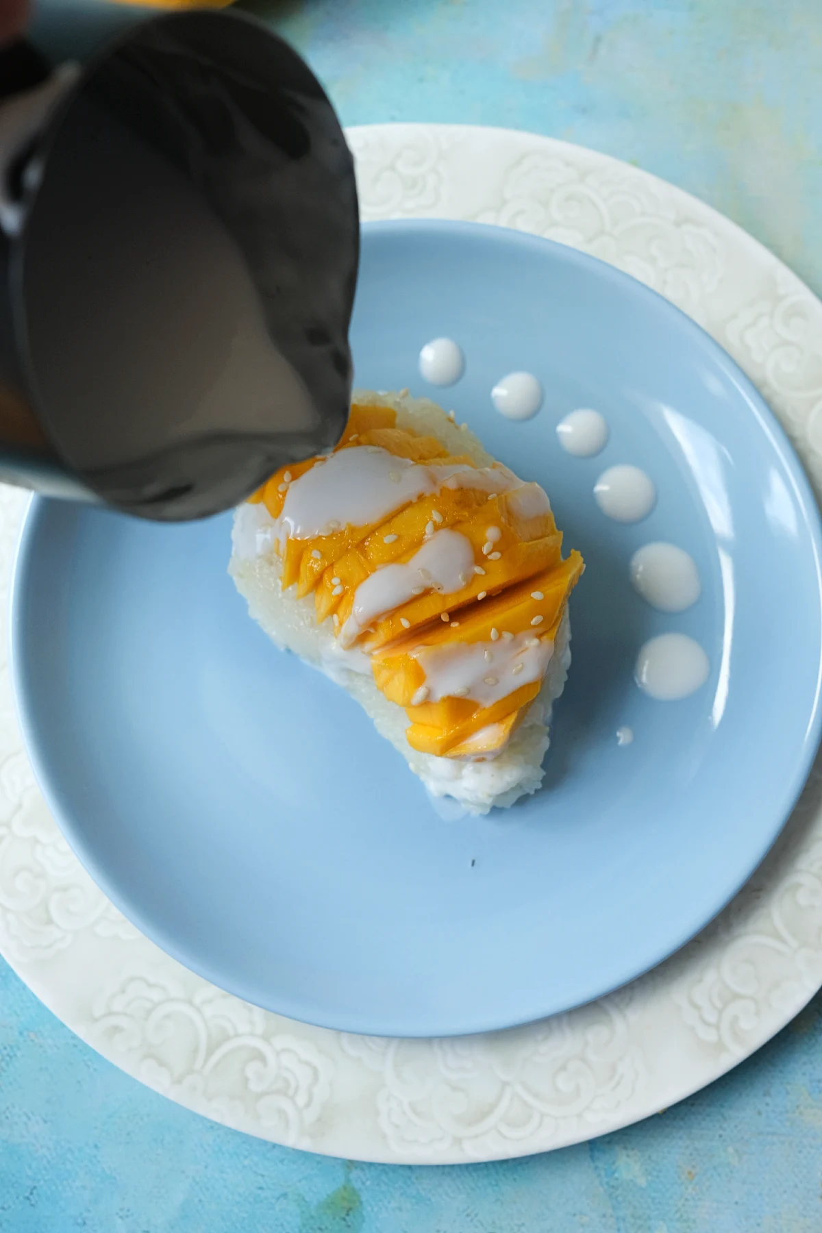 Coconut sauce being drizzled over mango sticky rice topped with fresh mango slices and sesame seeds.