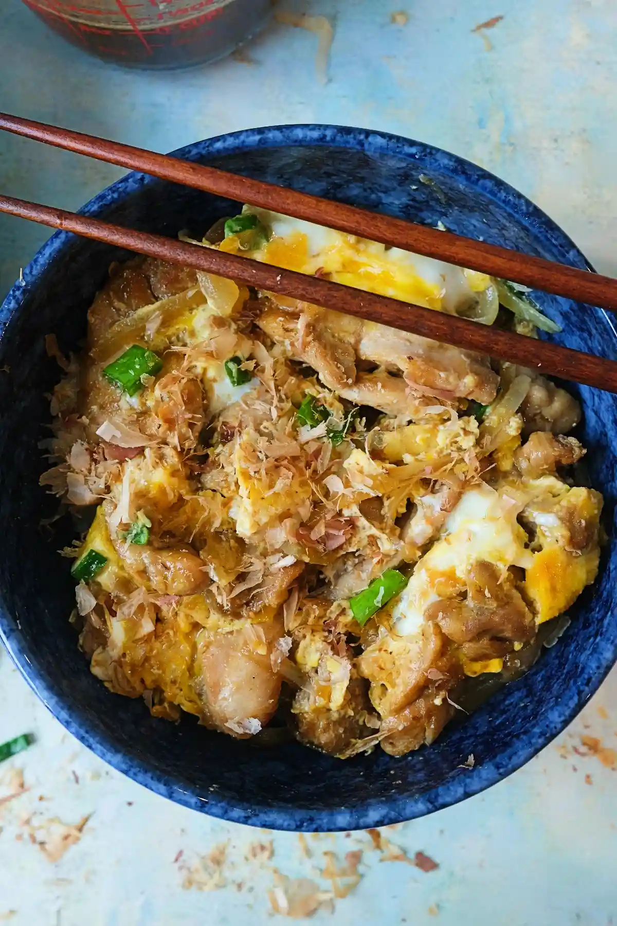 Close-up of a bowl of oyakodon with glossy chicken, soft scrambled eggs, green onions, and bonito flakes, with sauce visible on top.