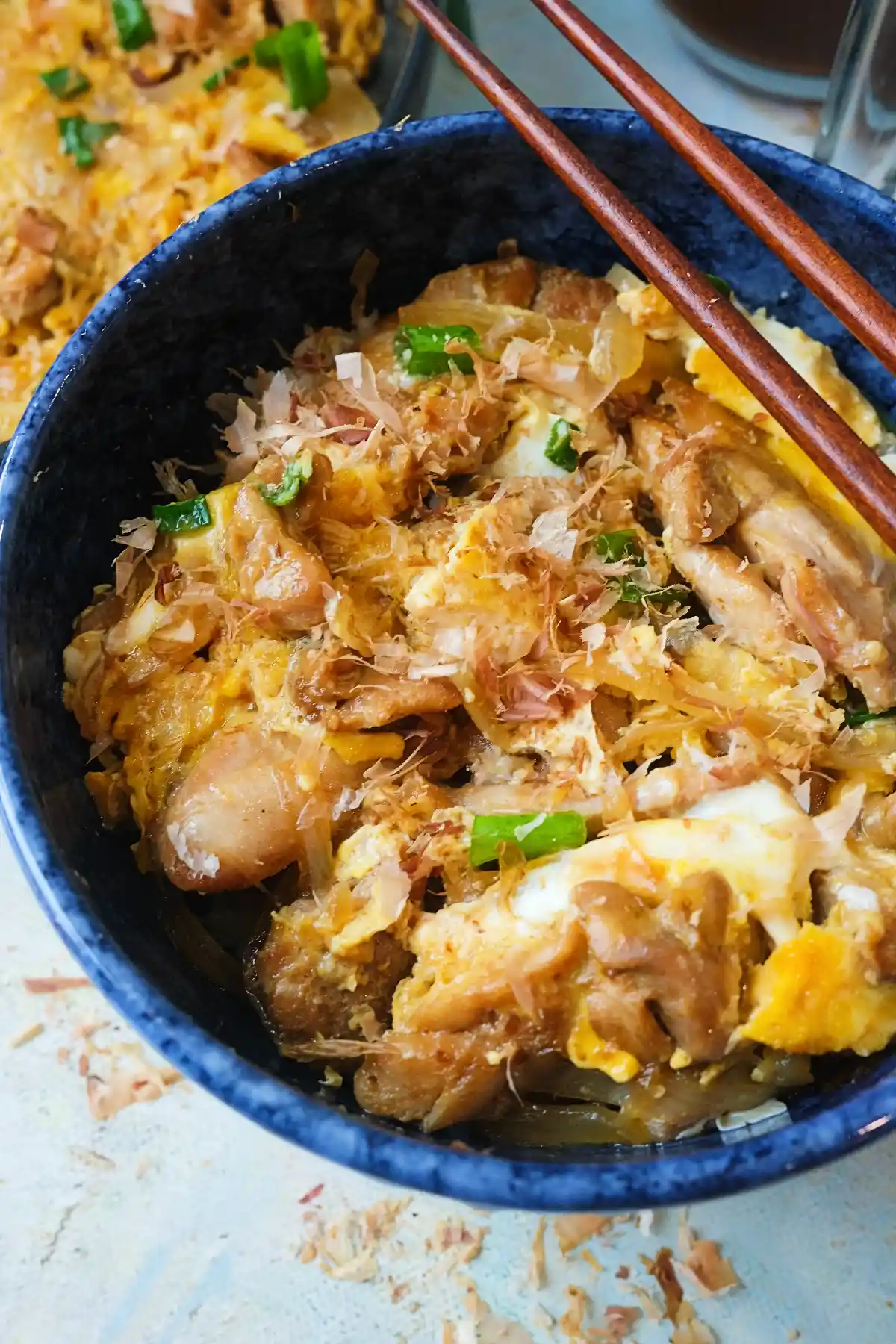 Close-up of oyakodon in a blue bowl topped with chicken, soft eggs, green onions, and bonito flakes with chopsticks resting on top.