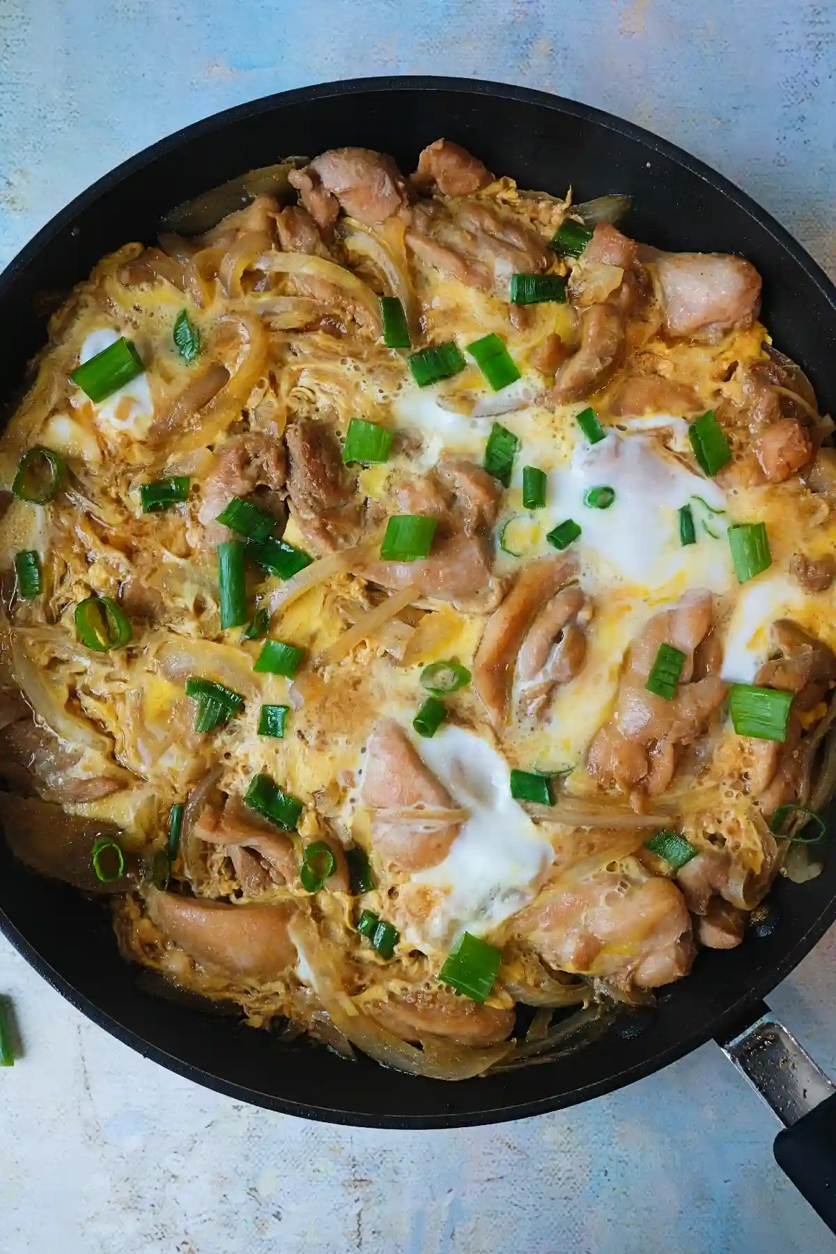 Oyakodon in a pan with chicken, onions, and partially set eggs, topped with chopped green onions.