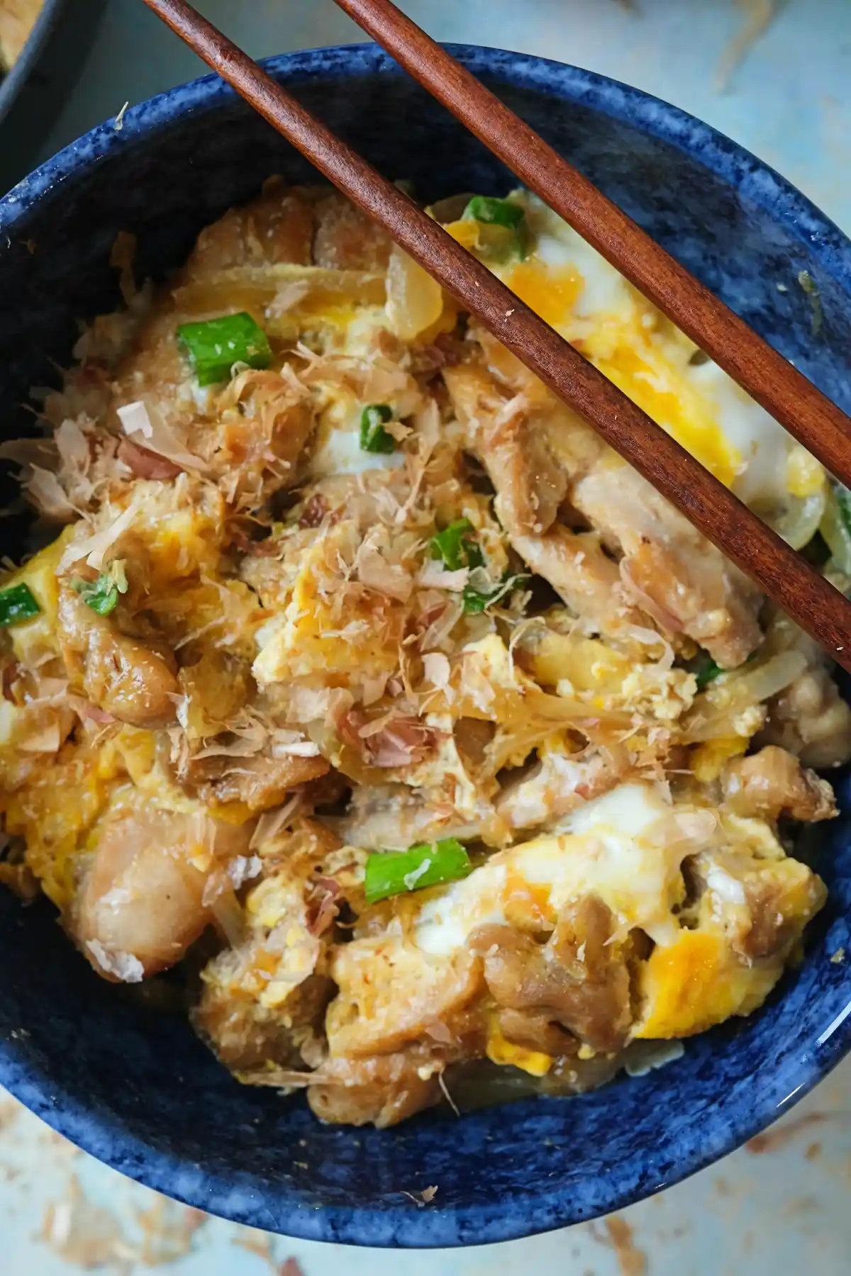 Overhead view of oyakodon in a bowl with tender chicken, softly set eggs, and green onions, with chopsticks placed across the bowl.