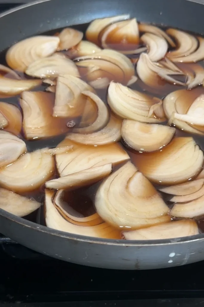 Sliced onions simmering in soy and dashi mixture in a pan.