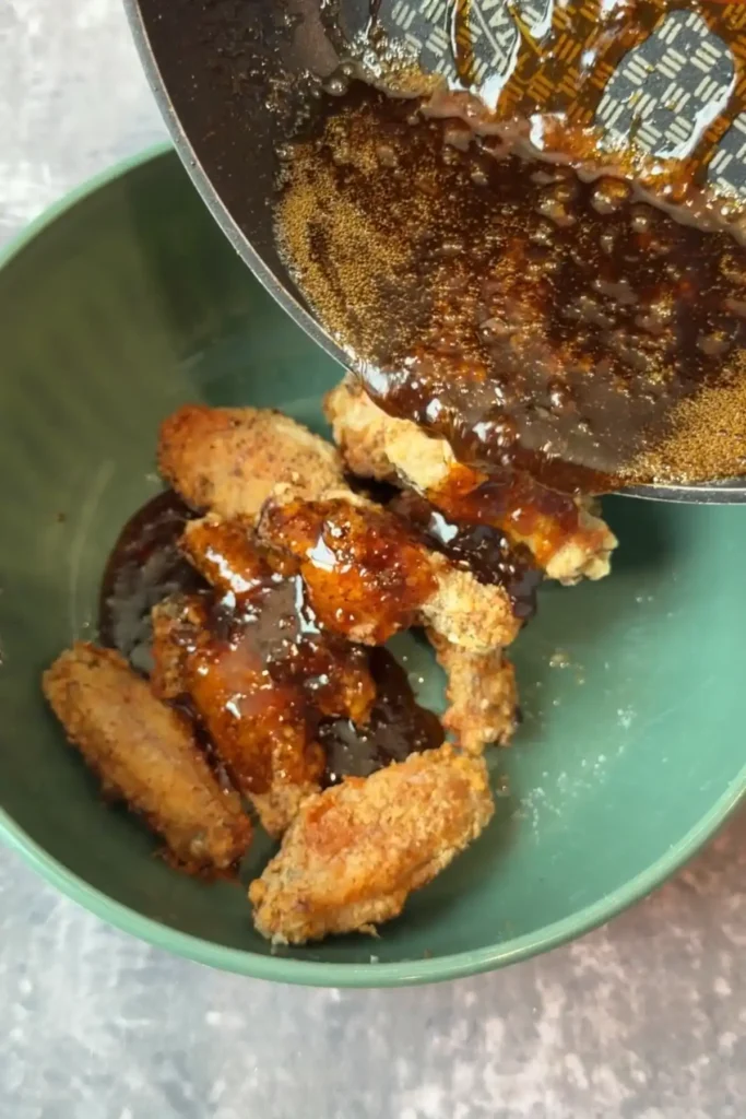Thick glossy soy garlic sauce pouring over crispy chicken wings in a mixing bowl.