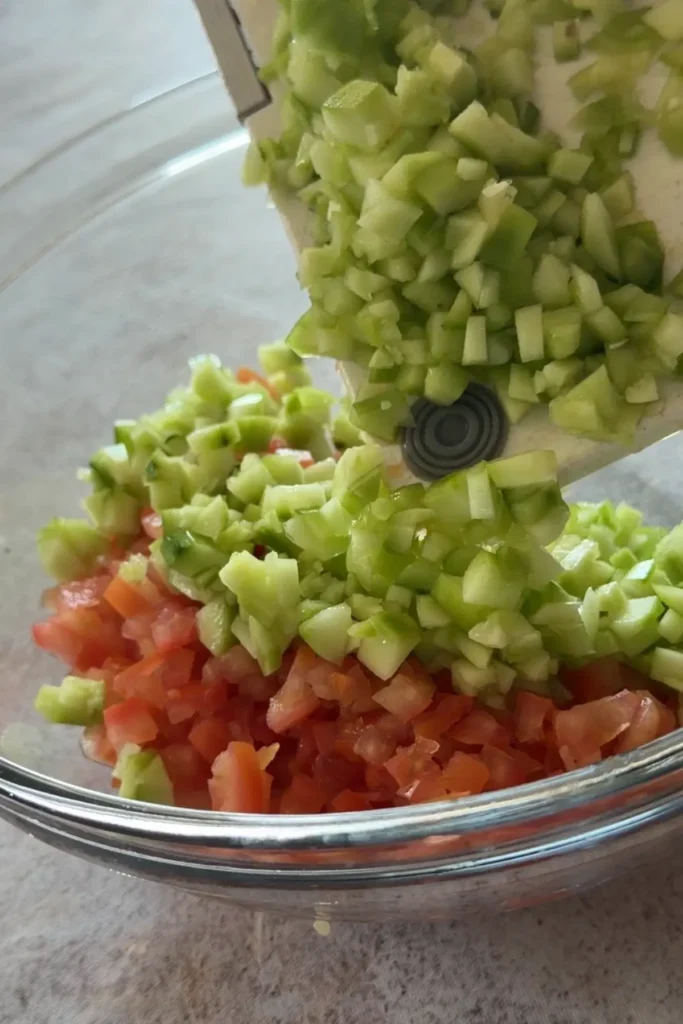 Diced cucumber being transferred from a cutting board into a glass mixing bowl.