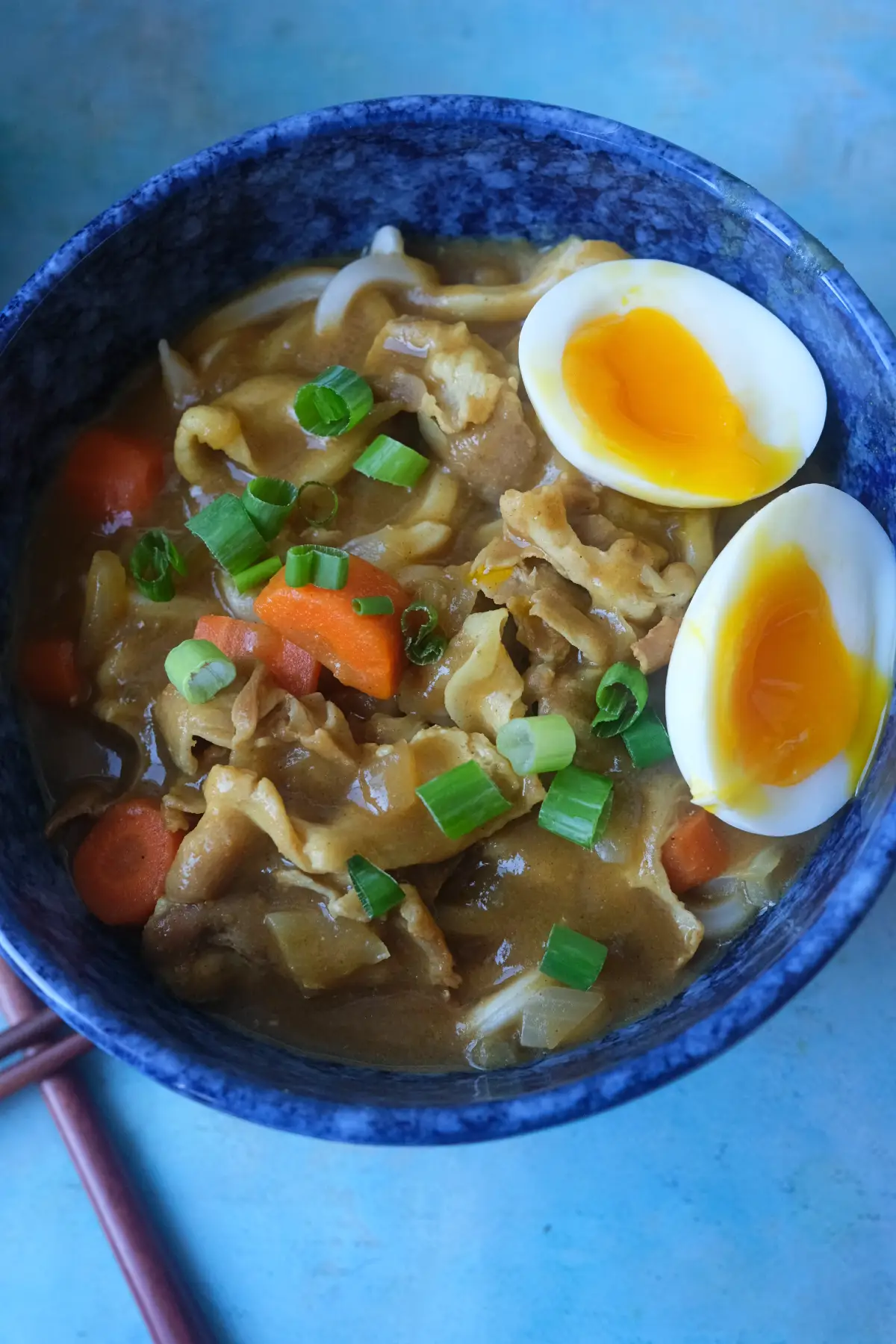 Japanese curry udon with pork belly, carrots, green onions, and a soft-boiled egg served in a blue bowl.