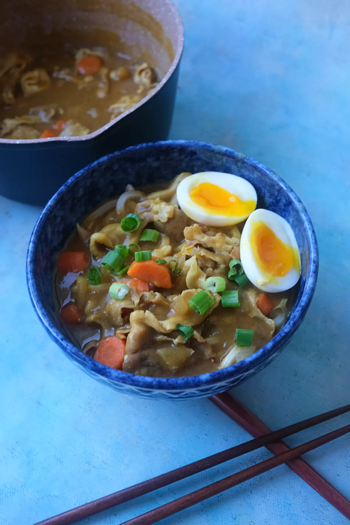 Bowl of Japanese curry udon with pork belly, carrots, green onions, and soft-boiled egg served beside the cooking pot.