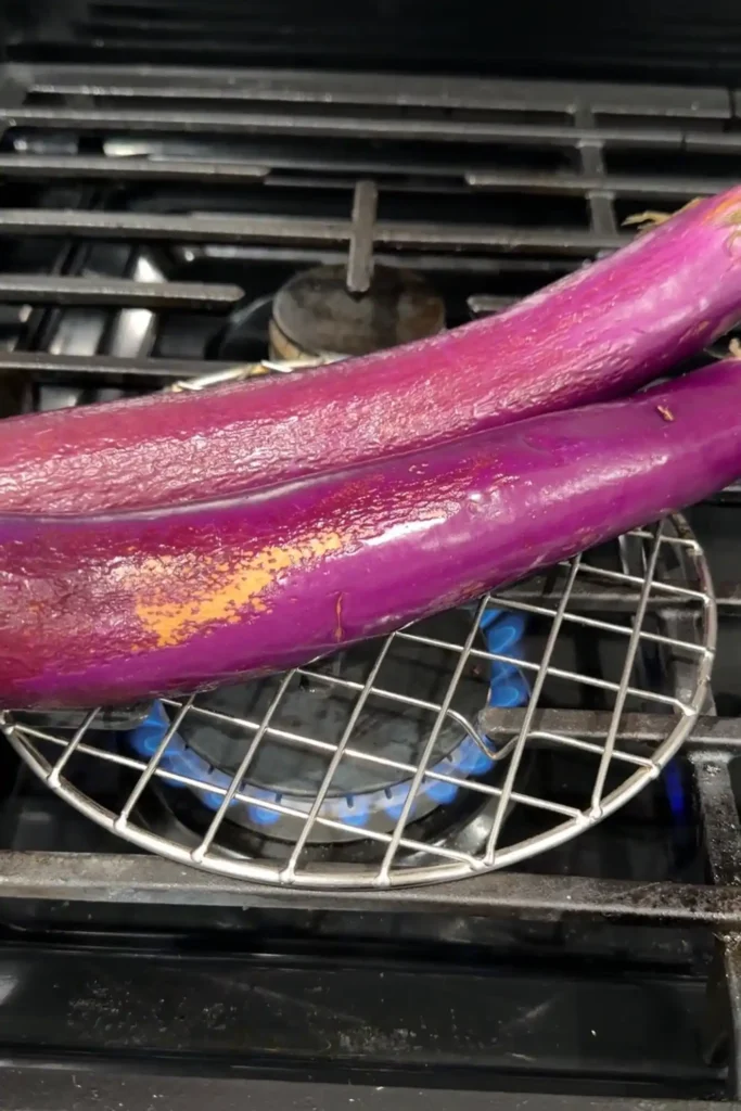 Long purple eggplants roasting over an open gas flame on a wire grilling rack.