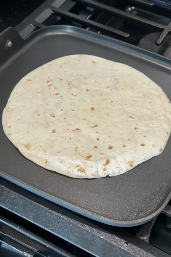 Large flour tortilla warming on a griddle before assembling ground beef quesadillas.