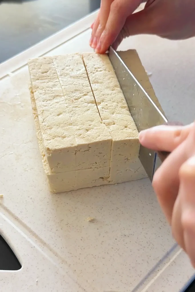 Extra-firm tofu being cut into cubes on a cutting board for tofu sisig.