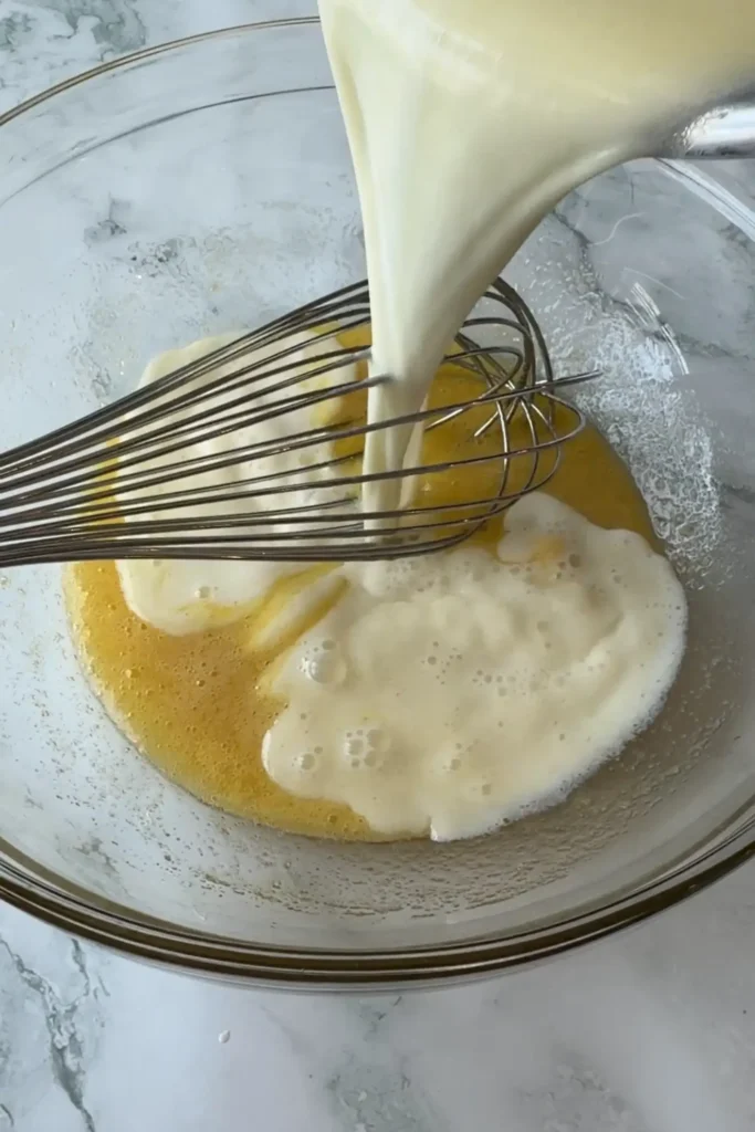 Warm milk and butter mixture being poured into the egg mixture while whisking.