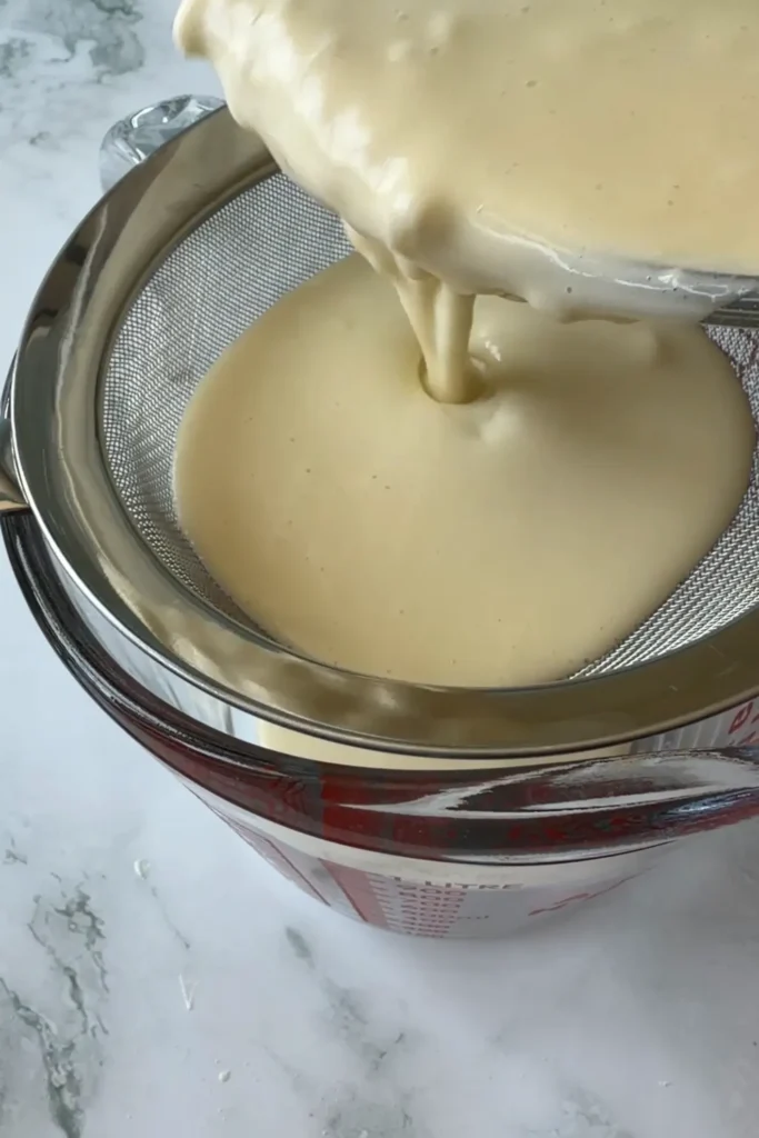 Butter tteok batter being poured through a fine-mesh sieve into a measuring cup.