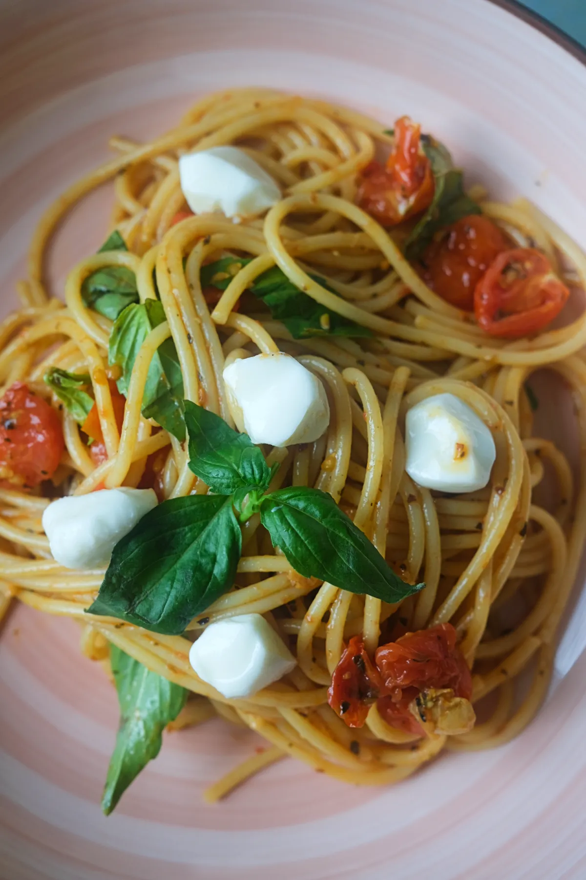 A serving of Caprese pasta with spaghetti, cherry tomatoes, fresh basil, and mozzarella pearls on a pink plate.