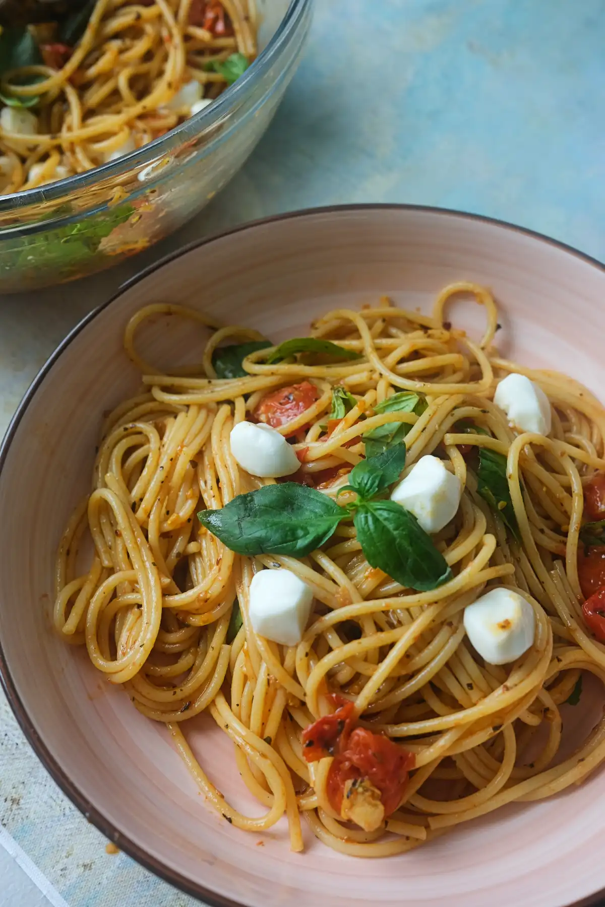 A bowl of Caprese pasta topped with fresh basil leaves and mozzarella pearls, with a glass mixing bowl of more pasta in the background.