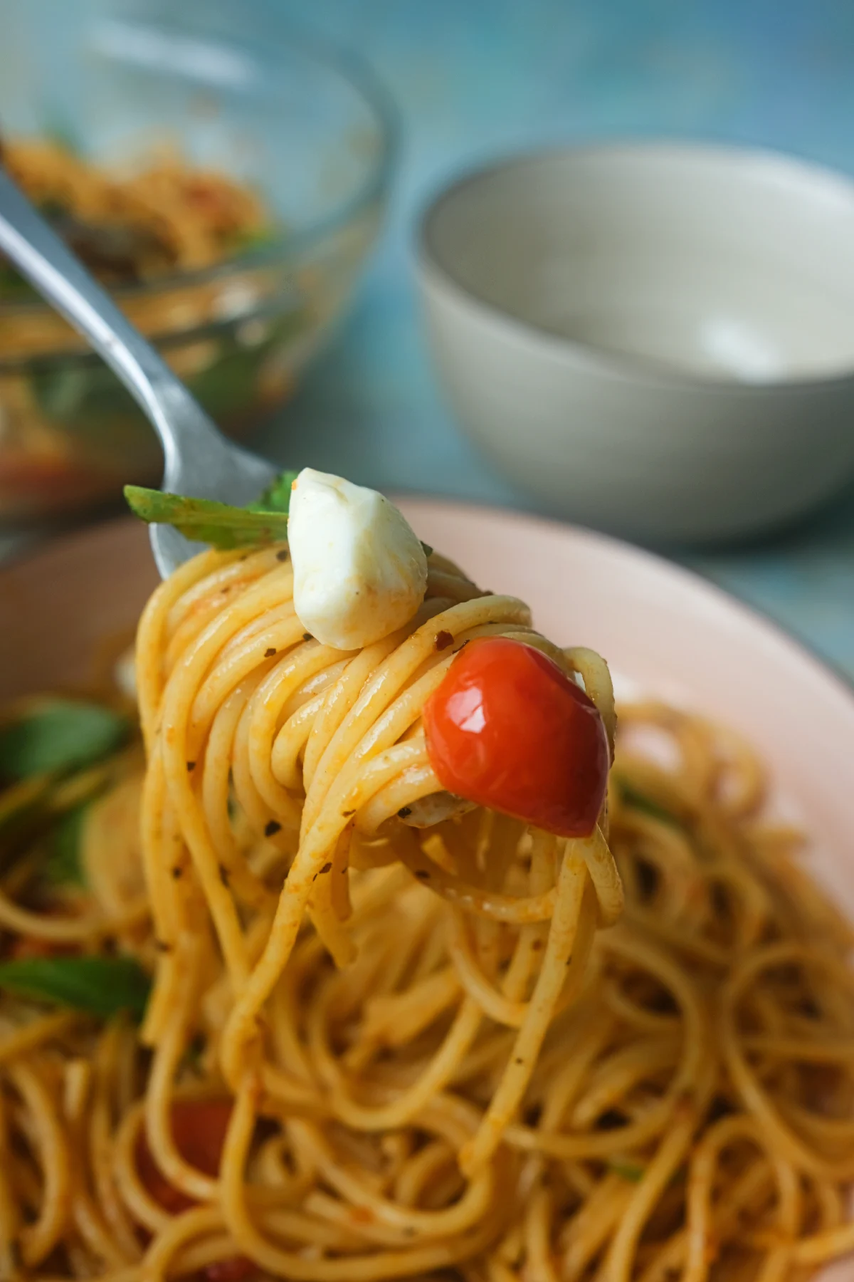 A forkful of spaghetti with a mozzarella pearl, blistered cherry tomato, and fresh basil leaf lifted above a bowl of Caprese pasta.