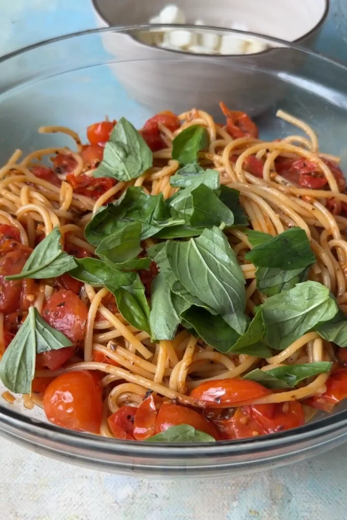 Fresh basil leaves added to the cooked spaghetti and blistered tomatoes in a glass mixing bowl.