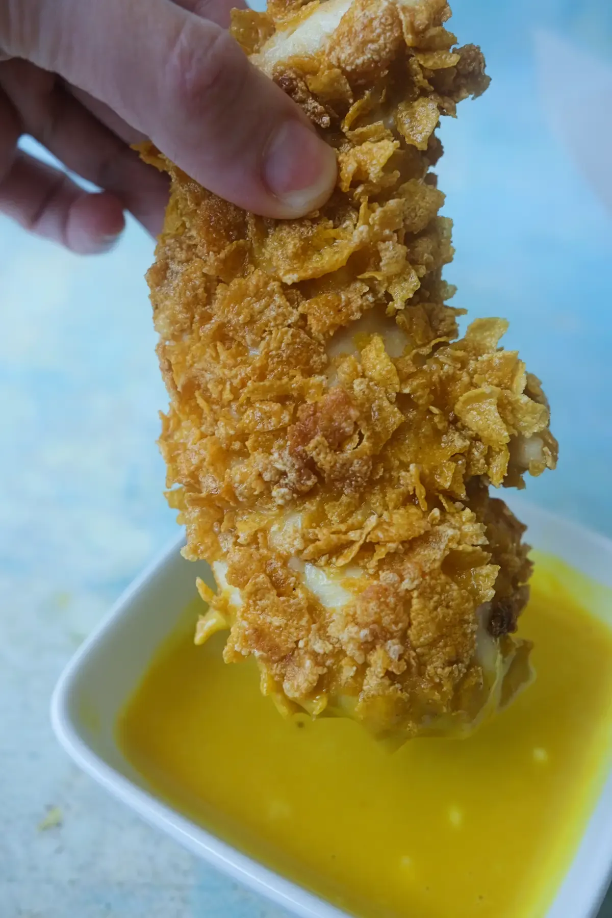 Hand dipping a crispy cornflake chicken tender into honey mustard sauce.