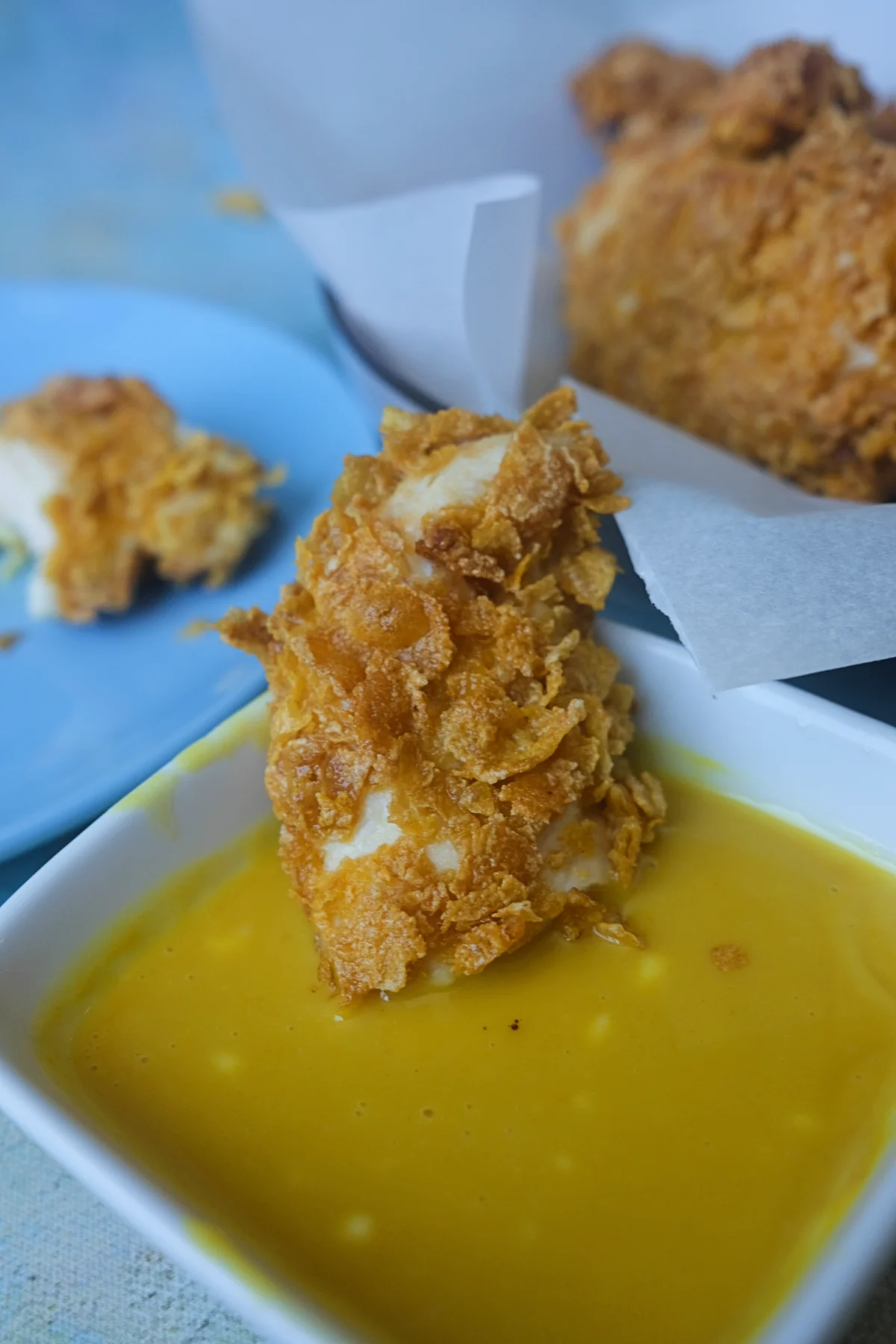 Crispy cornflake chicken tender dipped in honey mustard sauce with more tenders in the background.