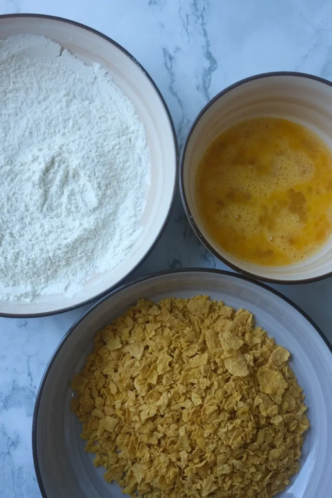 Three-bowl breading station with flour, beaten eggs, and crushed cornflakes.