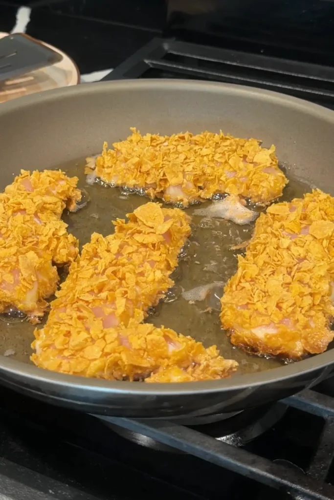 Cornflake-coated chicken tenders frying in a skillet with hot oil.