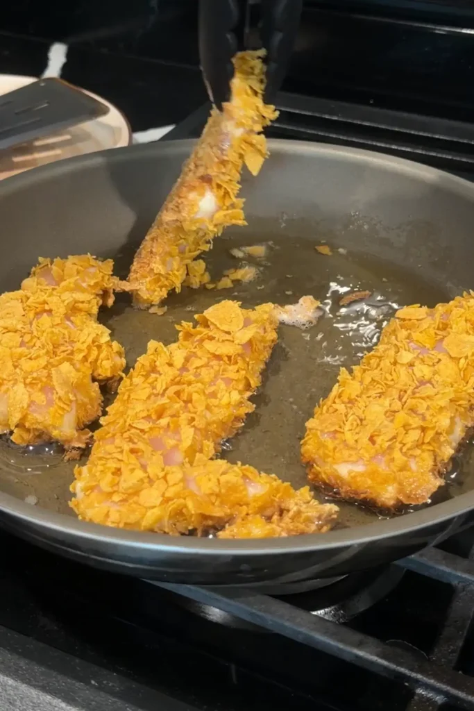 Tongs flipping a cornflake-coated chicken tender while frying in a skillet.