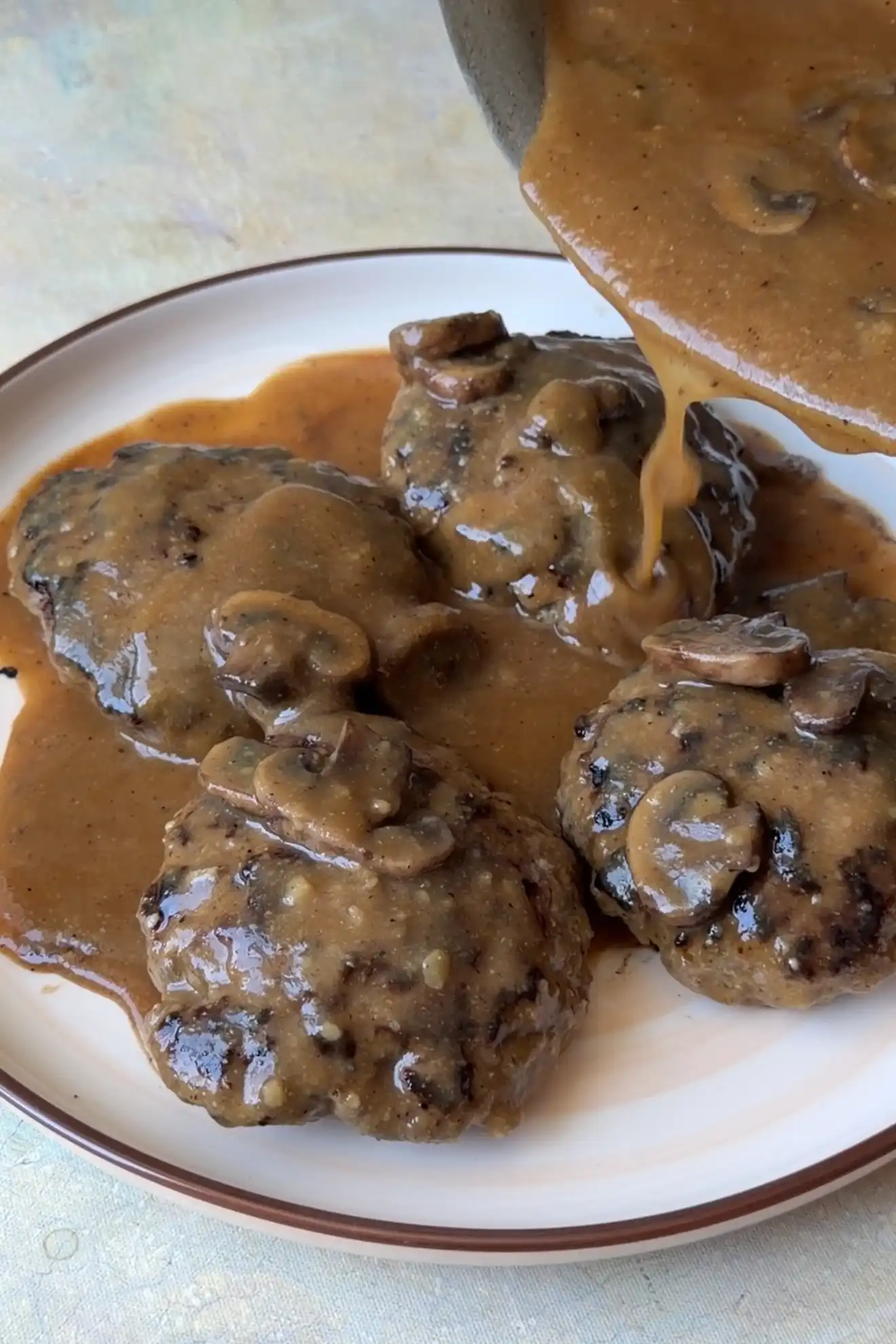 Mushroom gravy being poured over homemade hamburger steak patties on a serving plate.