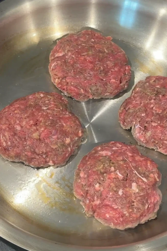 Homemade hamburger steak patties searing in a stainless steel skillet until browned.