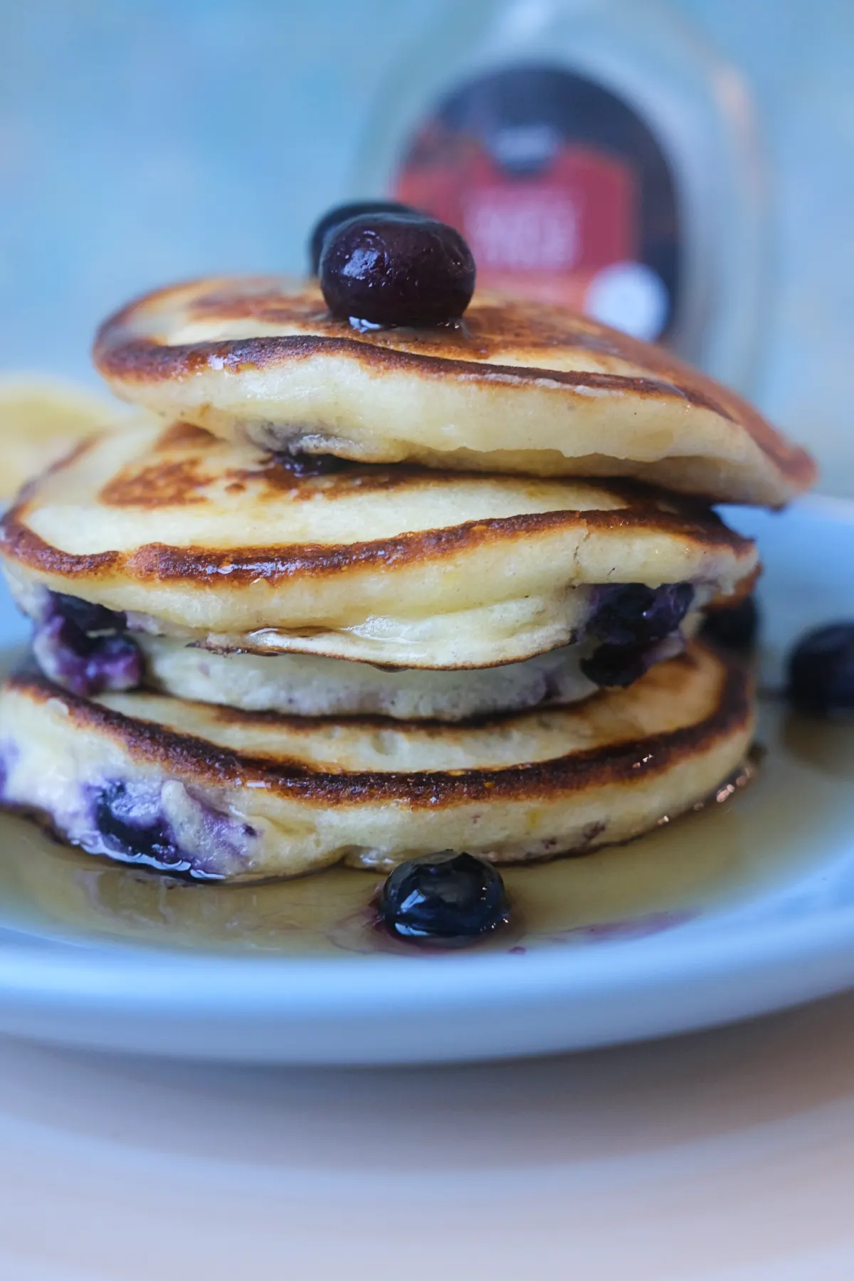 Lemon blueberry ricotta pancakes stacked on a plate with maple syrup, fresh blueberries, and a bottle of syrup in the background.