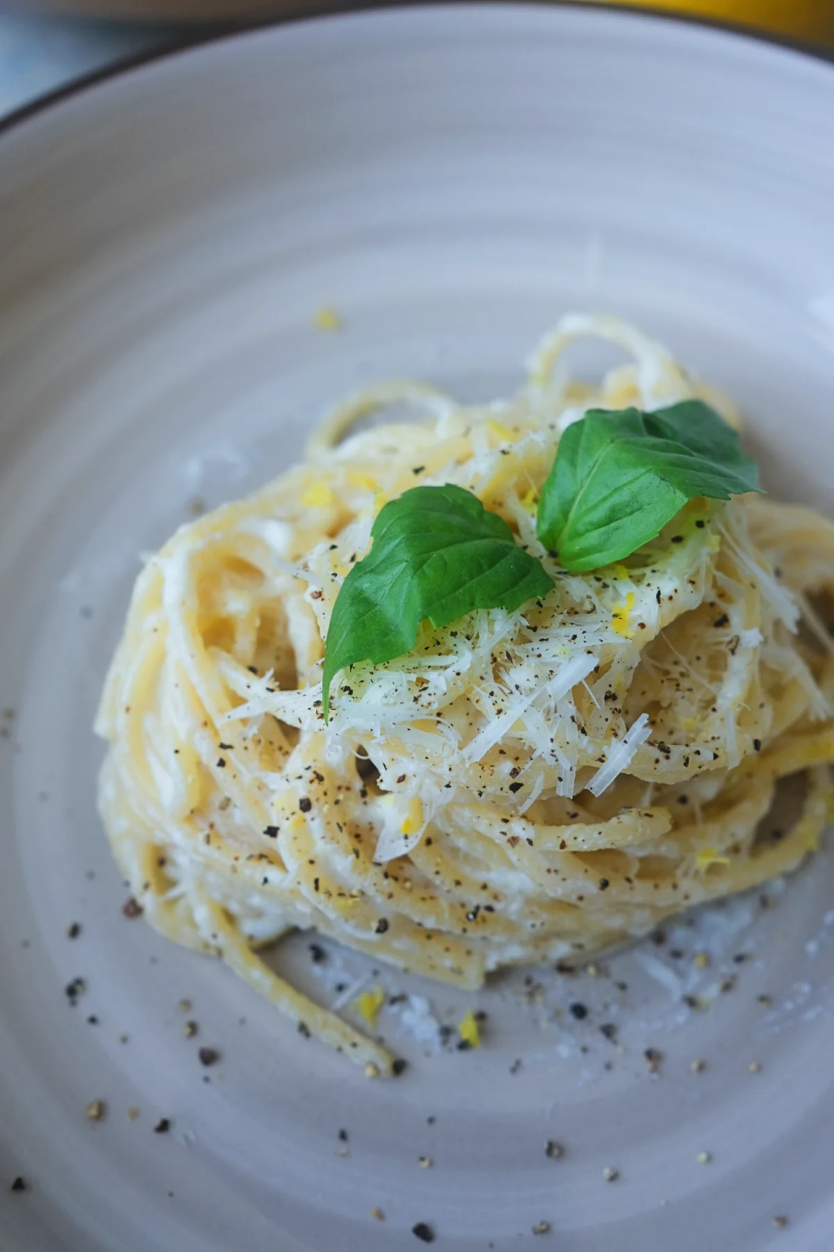 A plated serving of creamy lemon ricotta spaghetti garnished with grated Parmesan, basil, black pepper, and lemon zest, with bowls of ricotta and pasta blurred in the background.