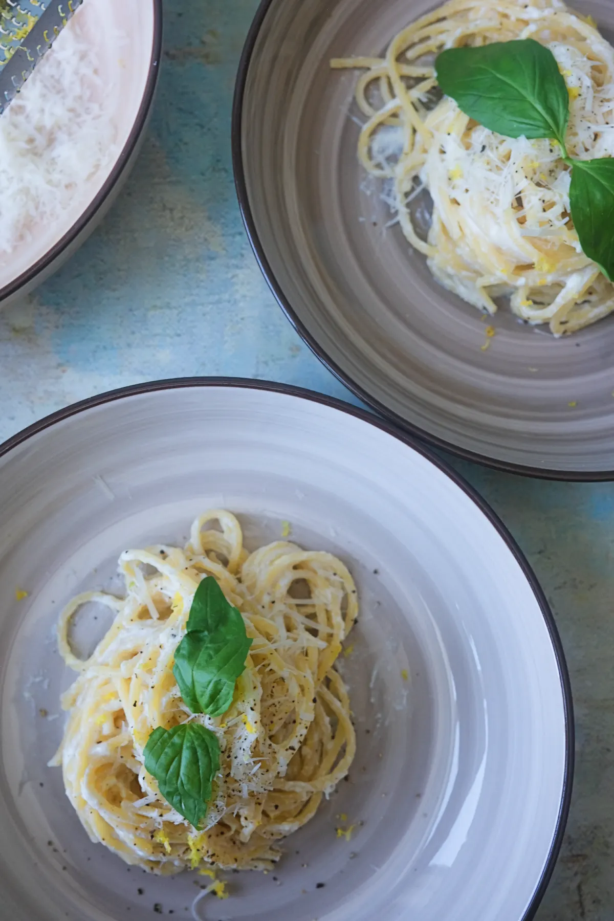 Overhead view of two plates of lemon ricotta pasta garnished with fresh basil, grated Parmesan, black pepper, and lemon zest beside a bowl of ricotta cheese and a microplane grater.